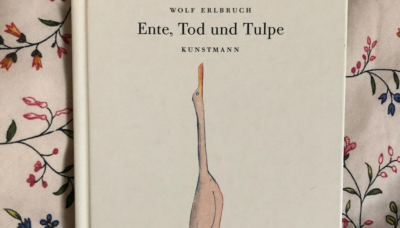Das Bilderbuch "Ente, Tod und Tulpe" von Wolf Erlbruch liegt auf einem geblümten Kissen. Auf dem Cover des Buches ist die Ente zu sehen, die nach oben schaut (sie sieht aus wie ein Gans: weiß und langer Hals).